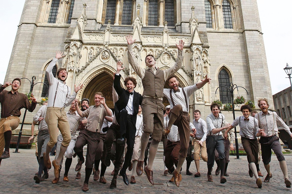 Choir leaping facing the camera with Truro Cathedral behind them
