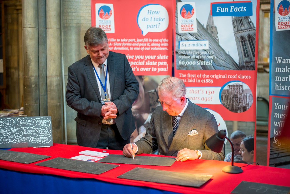 Kevin Bray standing whilst a seated Prince Charles sits at a table and signs a slate in support of Truro Cathedral's roof appeal. Banners for the roof appeal can be seen in the background.