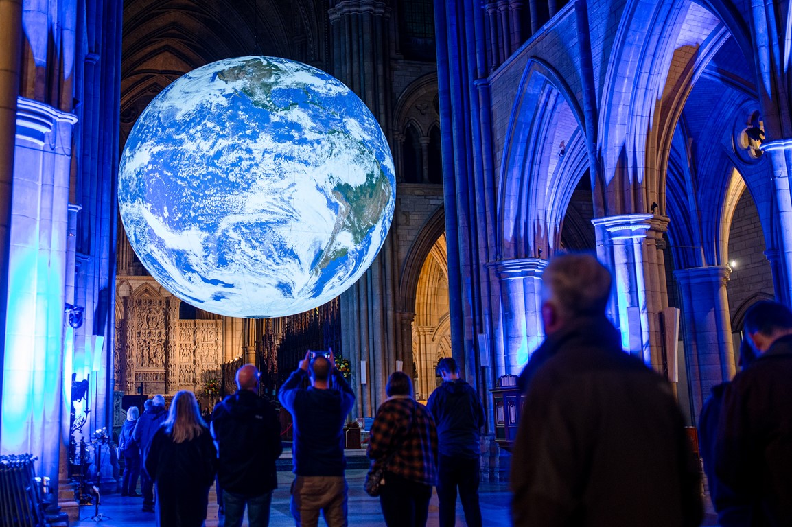 Illumnated Gaia globe suspended in Truro Cathedral, low level blue lighting, people gathered in foreground with backs to camera looking up at the art installation